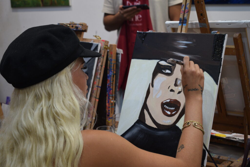 Persona con gorra negra y cabello rubio largo pintando un retrato en un lienzo durante un taller de arte, con detalles artísticos de un rostro en tonos oscuros y expresivos.