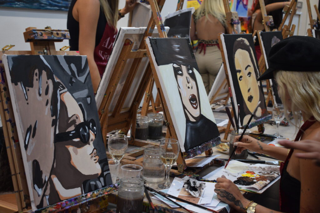 Varias personas participan en un taller de pintura, trabajando en retratos de estilo expresionista en lienzos. Se ven diferentes obras en proceso en los caballetes, con rostros pintados en tonos oscuros y contrastantes. Una persona, con gorra negra, pinta en primer plano mientras sostiene un pincel y paleta, con vasos de agua y pinceles alrededor.