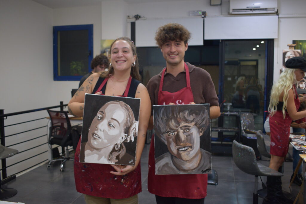 Dos personas, una mujer y un hombre, sostienen retratos pintados a mano en un taller de arte. Ambos llevan delantales rojos, y sonríen mostrando sus obras, una representación en tonos grises y marrones de rostros humanos.